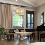 A modern dining area with a wooden table, black legs, and benches, illuminated by a contemporary pendant light. Large windows with beige curtains and a glass door provide natural light.